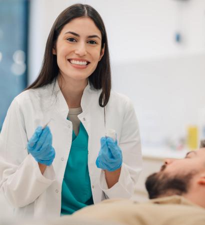 Dentist smiling and holding tools with patient lying down Smiling female dentist holding dental tools while male patient lies comfortably in the dentist chair, surrounded by bright, clean equipment in a modern dental surgery