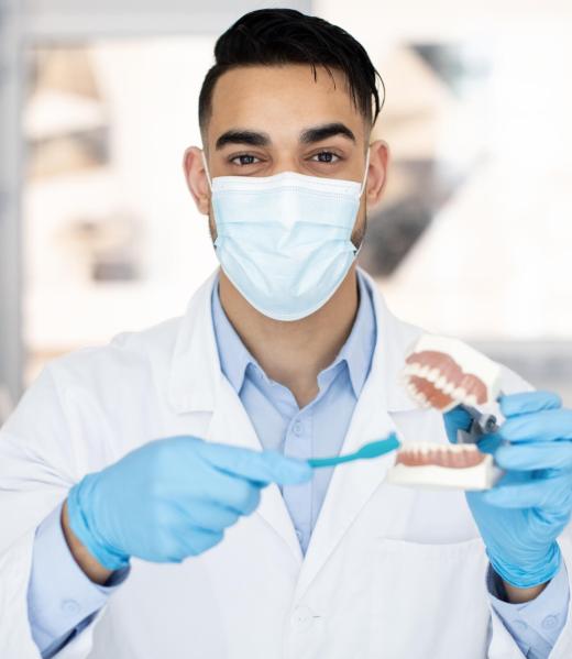 Oral and Dental Health. Arab Dentist Doctor Holding Jaw Model And Toothbrush Oral and Dental Health. Arab Dentist Doctor Holding Jaw Model And Toothbrush, Middle Eastern Stomatologist Wearing Medical Mask Demonstrating Right Way To Clean Teeth And Gums, Copy Space